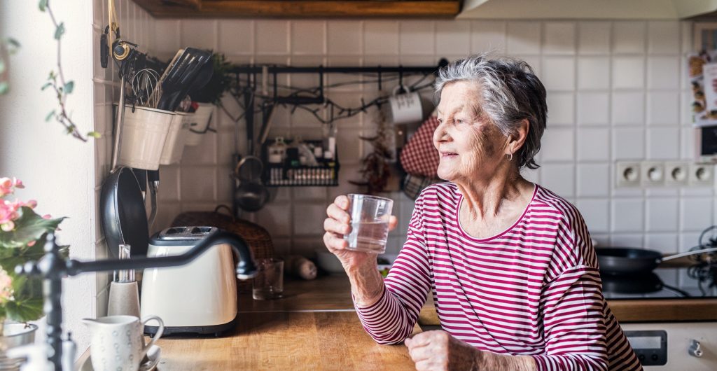 An elderly woman standing in the kitchen, holding a glass of water.