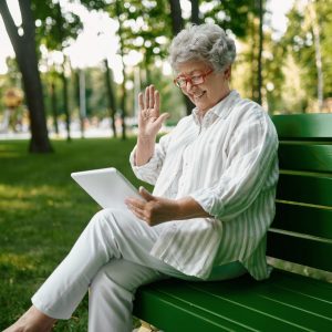 An elderly woman in glasses using laptop on bench